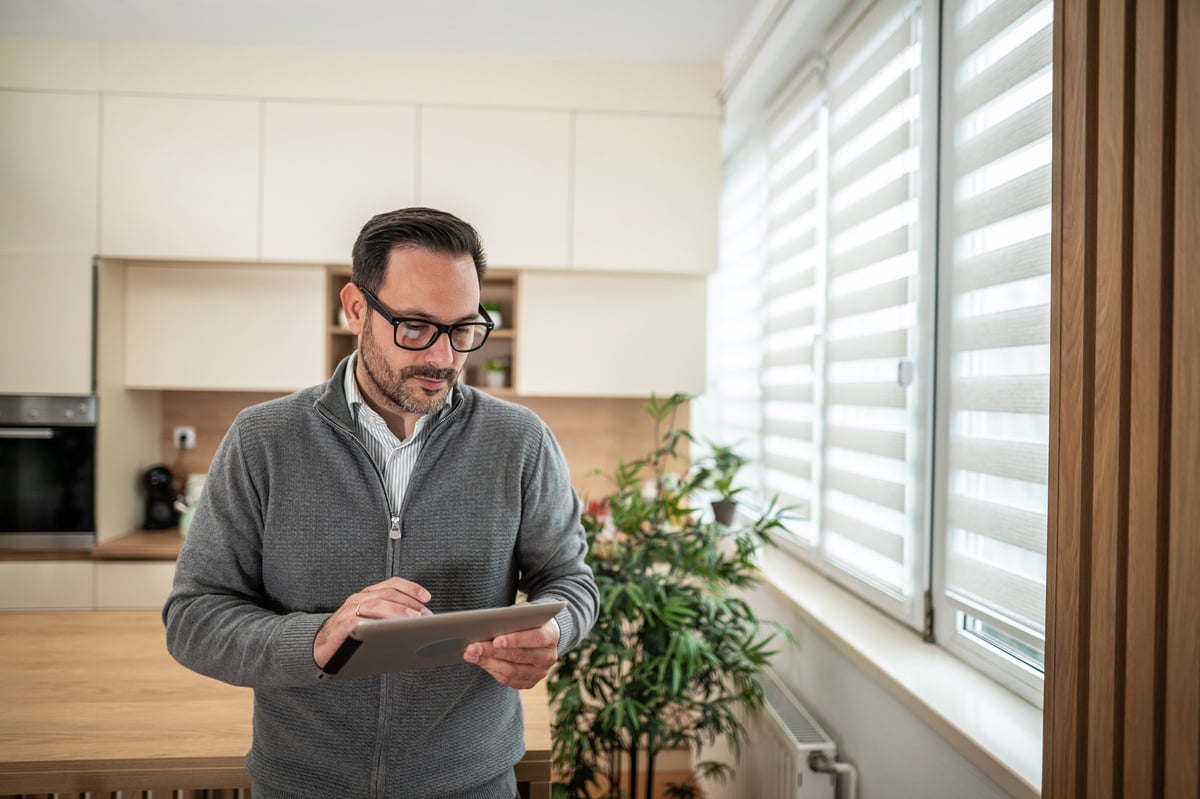 Man in casual clothes uses a tablet to control smart home features like roller blinds and lighting, checking automation settings in his modern, efficient apartment interior